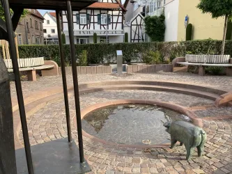 Ein kleiner Platz mit einem runden Brunnen in der Mitte. Ein bronzene Statue eines Schweins steht neben dem Brunnen, umgeben von Bänken und Pflanzen.