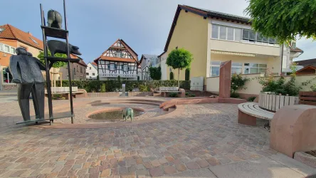 Ein schöner Platz mit einer runden Wasserstelle und mehreren Sitzbänken. Im Hintergrund sind traditionelle und moderne Gebäude zu sehen.
