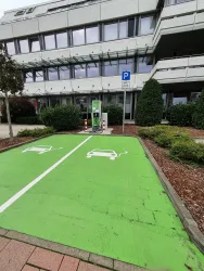 Eine Ladestation für Elektroautos befindet sich vor einem modernen Gebäude. Die Parkplätze sind grün markiert und zeigen Symbole für Elektrofahrzeuge.