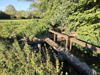 Ein kleiner Holzsteg führt über einen ruhigen Bach, umgeben von üppiger Pflanzenwelt. Im Hintergrund sind grüne Wiesen und Bäume sichtbar.