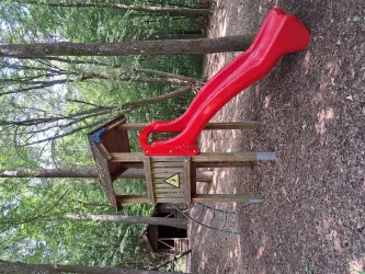 Eine Rutsche in einem Spielplatz aus Holz, umgeben von Bäumen. Der Spielplatz hat eine blaue Dachfläche und eine Sicherheitswarnung.