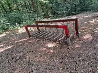 Ein Holzgestell steht im Wald, umgeben von Laub und Bäumen. Die Szene zeigt eine ruhige und natürliche Umgebung.