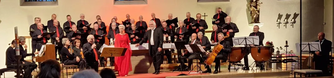 Ein Chor und ein Orchester treten auf einer Bühne auf. Die Sänger sind in schwarzen Roben gekleidet, während eine Solistin in einem roten Kleid hervorsticht.