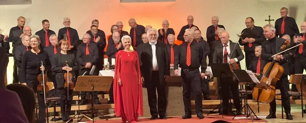 Ein vollständiger Chor steht auf der Bühne, während sie singen. Im Vordergrund stehen Künstler in eleganter Kleidung, eine Person trägt ein rotes Kleid.