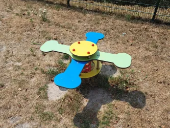 Ein buntes Spielgerät mit einer runden Plattform und vier Armen steht auf trockenem Gras. Es ist ein sonniger Tag in einem Spielplatzbereich.