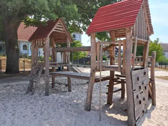 Ein Spielplatz mit zwei Holzspielhäuschen und einer Rutsche. Der Boden ist mit Sand bedeckt und es gibt einen großen Baum im Hintergrund.