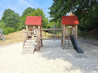 Ein Spielplatz mit zwei Spieltürmen aus Holz und roten Dächern. Auf der einen Seite gibt es eine Rutsche und den Sandboden umgibt eine grüne Naturlandschaft.