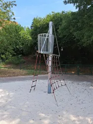 Ein Spielplatzgerät in Form einer Kletterstruktur steht auf einem sandigen Boden. Im Hintergrund sind grüne Bäume sichtbar.
