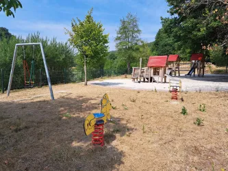 Ein Spielplatz mit einer Schaukel und einem Spielhaus steht in einer grünen Umgebung. Die Fläche ist trocken mit wenigen Pflanzen.