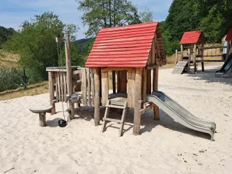 Ein Spielplatz mit einer hölzernen Spieleinheit und einer Rutsche. Der Boden ist mit Sand bedeckt und es gibt Bäume im Hintergrund.