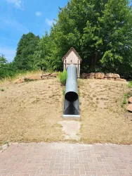 Eine Rutsche aus Metall, die von einem kleinen Holzhaus auf einem Hügel herunterführt. Im Hintergrund sind Bäume und eine grüne Wiese zu sehen.