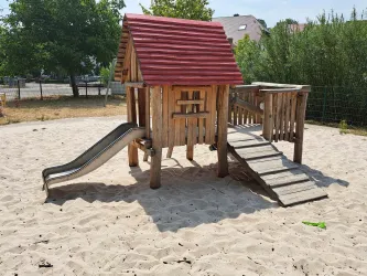 Ein Holzspielplatz mit einem roten Dach, einer Rutsche und einer Brücke. Der Spielplatz steht auf einem Sandbereich und ist von Bäumen umgeben.