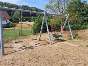 Ein Spielplatz mit zwei herkömmlichen Schaukeln und einer großen Nestschaukel. Im Hintergrund sind Bäume und eine Wiese zu sehen.