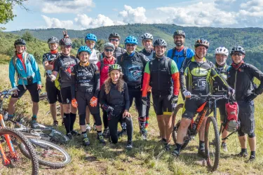 Eine Gruppe von Mountainbikern steht fröhlich auf einer Wiese mit Bergen im Hintergrund. Die Teilnehmer tragen bunte Radbekleidung und Helme.