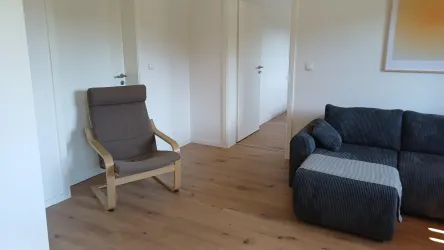 Ein heller Raum mit einem bequemen Sessel und einem grauen Sofa. Der Boden ist aus Holz und die Wände sind weiß gestrichen.
