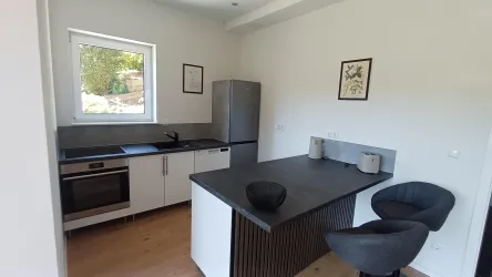 Eine moderne Küche mit weißen Schränken und einem grauen Küchenblock. Der Raum ist hell und hat große Fenster, die natürliches Licht hereinlassen.
