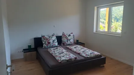 Ein modernes Schlafzimmer mit einem Doppelbett und bunten Kissen. Große Fenster sorgen für viel Tageslicht und Blick auf die Natur.