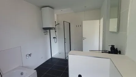 Ein modernes Badezimmer mit einer Dusche und einem Waschbecken. Die Wände sind hell, und der Boden ist mit schwarzen Fliesen ausgelegt.