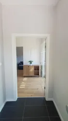 Ein heller Flur mit einem offenen Durchgang. Im Hintergrund ist ein Wohnzimmer mit einem Holzmöbelstück und einer Pflanze zu sehen.