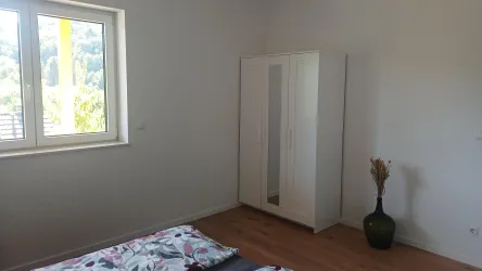Ein einfaches Schlafzimmer mit einem Bett, einem Kleiderschrank und einem Fenster. Die Wände sind hell, und der Boden ist aus Holz.