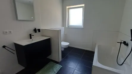 Ein modernes Badezimmer mit einem Waschbecken und einer Badewanne. Der Raum ist hell und geräumig, mit schwarzen Fliesen und einem Fenster.