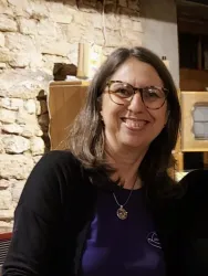 Eine lächelnde Frau mit langen, braunen Haaren und Brille sitzt vor einer Steinmauer. Sie trägt ein schwarzes Oberteil und eine Kette.