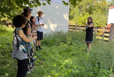 Eine Gruppe von fünf Personen steht in einem grünen Garten. Eine Frau erklärt etwas, während die anderen aufmerksam zuzuhören.