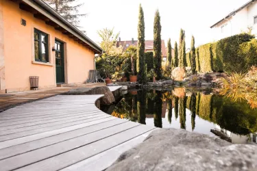 Ein ruhiger Garten mit einem Teich und einem Holzsteg. Im Hintergrund sind hohe Zypressen und ein gemütliches Gebäude zu sehen.
