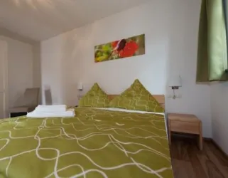 Ein gemütliches Schlafzimmer mit einem Doppelbett und grüner Bettwäsche. An der Wand hängt ein großes farbiges Bild und ein Fenster mit grünem Vorhang sorgt für Tageslicht.