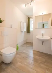 Ein modernes Badezimmer mit hellen Fliesen und einer sauberen Dusche. Es gibt eine Toilette und ein Waschbecken sowie eine helle Wandfarbe.