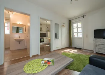 Ein modernes Wohnzimmer mit einer gemütlichen Couch und einem grünen Teppich. Der offene Raum führt in eine Küche und einen Flur mit natürlichem Licht.