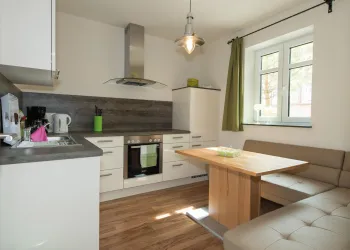Eine moderne Küche mit weißen Schränken und Holzfußboden. Ein Esstisch aus Holz und ein gemütlicher Sitzbereich sind ebenfalls vorhanden.