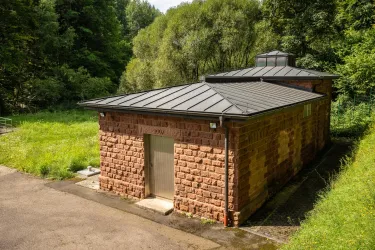 Ein modernes Gebäude aus roten Steinen in einer grünen Umgebung. Es hat ein flaches, schwarzes Dach und ist von Bäumen umgeben.