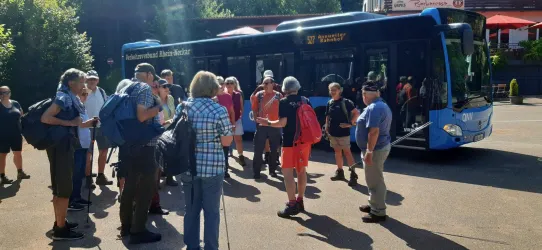 Eine Gruppe von Wanderern versammelt sich vor einem blauen Bus. Im Hintergrund sind Bäume und ein sonniger Himmel zu sehen.