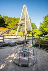 Ein Kind spielt auf einem Karussell in einem Spielplatz. Im Hintergrund steht eine große Rutsche unter klarem Himmel.
