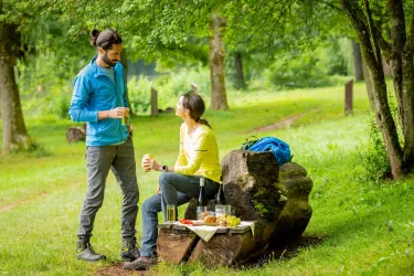 Ein Paar entspannt im Wald auf einem Baumstamm. Sie genießen Getränke und Snacks inmitten der Natur.