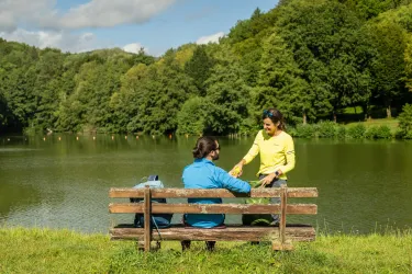 Zwei Personen sitzen auf einer Bank am Ufer eines ruhigen Sees. Im Hintergrund sind grüne Bäume und ein blauer Himmel zu sehen.