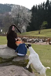 Ein Kind sitzt auf einem Stein und schaut zu einer Person im Hasenkostüm, die Futter hält. Im Hintergrund sind Ziegen und eine grüne Landschaft sichtbar.