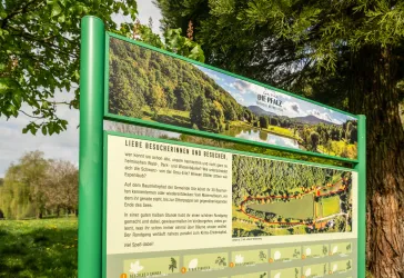 Ein Informationsschild über die Pfalz mit Bildern von der Landschaft. Die Grünflächen und der Fluss sind sichtbar, umgeben von Bäumen.