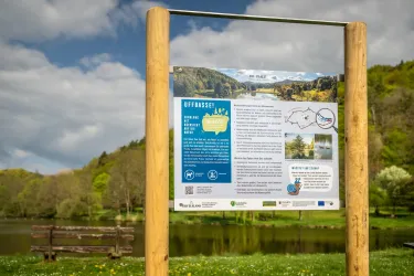 Ein Informationsschild am Ufer eines ruhigen Gewässers. Im Hintergrund sind Bäume und ein blauer Himmel mit wenigen Wolken zu sehen.