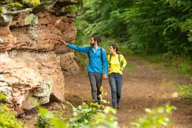 Zwei Wanderer gehen auf einem Waldweg und betrachten eine Steinformation. Die Umgebung ist grünen und friedlich.