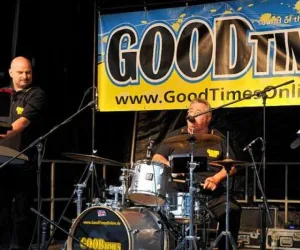 Eine Band spielt auf einer Bühne mit einer großen "GOODTIME"-Banner im Hintergrund. Der Schlagzeuger und ein Keyboarder sind aktiv in die Darbietung eingebunden.
