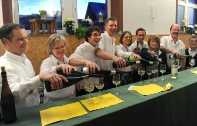 Eine Gruppe von sechs Personen in weißer Kleidung steht hinter einem Tisch und schenkt Wein in Gläser ein. Im Hintergrund sind einige Pflanzen und Fenster sichtbar.