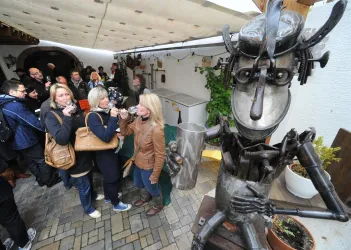 Eine belebte Veranstaltung mit vielen Menschen in einem Innenraum. Im Vordergrund steht eine große Statue aus Metall, die eine figurative Skulptur darstellt.