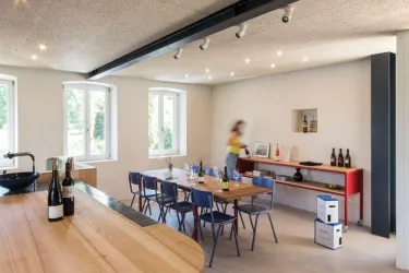 Ein stilvolles, modernes Esszimmer mit einem großen Tisch und blauen Stühlen. Im Hintergrund sind Flaschen und dekorative Elemente auf einem Regalsystem sichtbar.