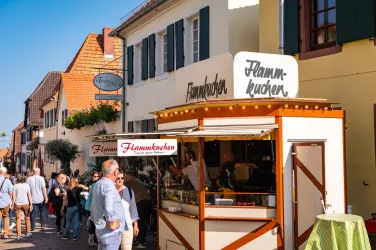 Ein lebhafter Marktstand verkauft Flammkuchen. Im Hintergrund sind Menschen und traditionelle Häuser zu sehen.