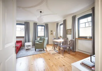 Ein stilvoll eingerichtetes Wohnzimmer mit einem roten Sofa, einem grünen Sessel und einem Esstisch. Große Fenster lassen viel Licht hereinfloßen und die Holzböden verleihen dem Raum Wärme.