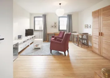 Ein einladendes Wohnzimmer mit zwei roten Sesseln und einem hellen Holzboden. Es gibt einen Tisch, einen Fernseher und große Fenster, die viel Licht hereinlassen.