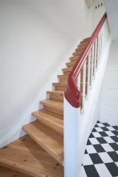 Eine helle Treppe aus Holz führt nach oben. Der Handlauf ist rot und der Boden hat ein schachbrettartiges Muster in Schwarz und Weiß.