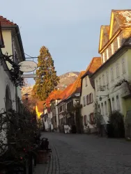 Eine ruhige Gasse mit historischen Gebäuden und gepflastertem Boden. Im Hintergrund sind Berge und ein klarer Himmel zu sehen.